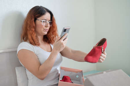 A frustrated woman unpacks an online purchase and takes a photograph of the shoe on a smartphone. The girl received the wrong packageの写真素材