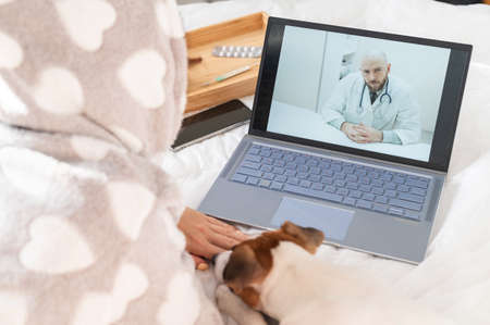 A sick woman lies in bed and talks with a male doctor on a laptop. Remote reception with a therapistの写真素材
