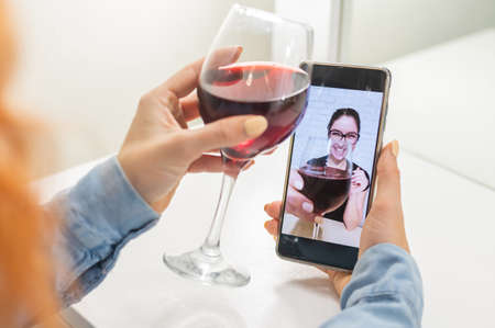 The women talk over video communication on the phone and drink red wine. The quarantined girl is not...