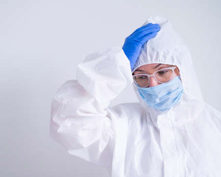 A woman in a protective suit and a medical mask is holding her head. Doctor virologist with problems at workの写真素材