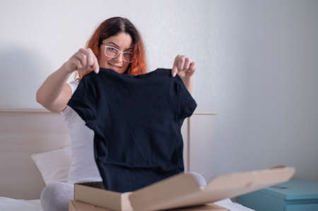 Woman unpacks online purchase of a T-shirt while sitting in bed. Home deliveryの写真素材