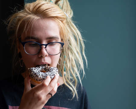 An unusual girl with dreadlocks on her head and a piercing eats a donut in a cafe. A young woman in glasses is having breakfast against a gray wall. Eating habits. Insulin.の写真素材