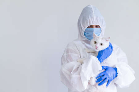 A woman in a protective suit with gloves and a mask is holding a white cat. Doctor tests pets for...