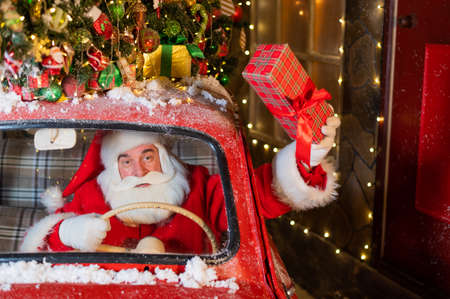 Santa Claus delivers presents behind the wheel of a red car and waves a gift box out of the windowの写真素材