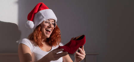 An excited woman in a Santa Claus hat takes red shoes from a cardboard box. The girl unpacks the delivered order for Christmas. Online shopping at homeの写真素材