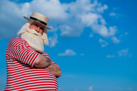 Elderly gray-haired man with a beard in a striped bathing suit and hat posing on the beach. Senior citizen on vacation by the lake.の写真素材
