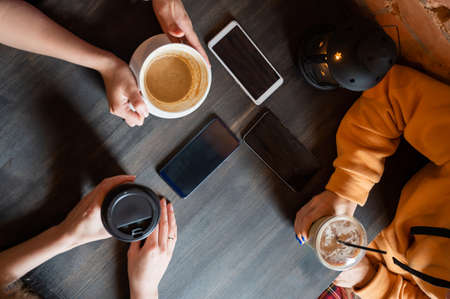 Top view of female hands with smartphones and cups of coffee on a table in a cafe. Three girlfriends drinking coffee.の写真素材