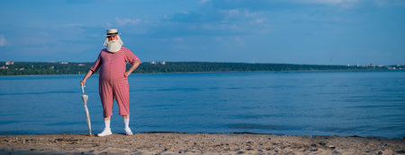 An elderly gray-haired man in a classic bathing suit and hat stands on the beach with an umbrella and waves his hand in greeting. Retired with a beard on vacation. Wide screenの写真素材