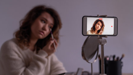 Young caucasian woman records video of online makeup training on a smartphone. Video blogger girl applies eye shadowの写真素材