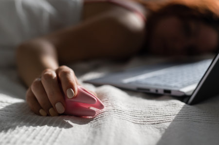 A woman lies in bed holding a clitoral vibrator and watching porn on a laptop. The girl has sex onlineの写真素材