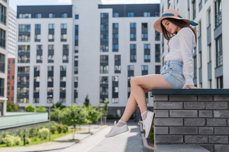Beautiful young woman in shorts and a hat posing while sitting on a stone bench. A full-length portrait of a Caucasian girl on a hot summer day.の写真素材
