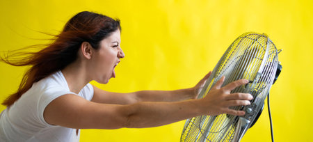 Emotional red-haired woman is cooled off standing over a large electric fan on a yellow background. Girl with hair developing in the wind. Device for cooling the air. Widescreenの写真素材