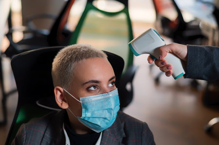 Business woman in medical mask has body temperature measured with electronic thermometer at workplace in office.の写真素材