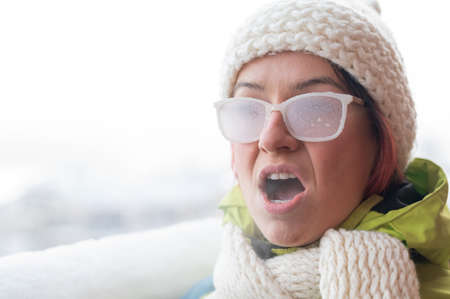 Portrait of a red-haired Caucasian woman in a white knitted hat and icy glasses outside in the frost