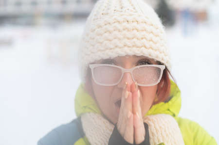 Portrait of a woman in glasses covered with hoarfrost. The girl is freezing and forgot gloves in very cold weather and blows on her bare handsの写真素材