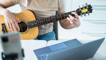 A woman sits in the kitchen during a remote acoustic guitar lesson. A girl learns to play the guitar and watches educational videos on a laptopの写真素材