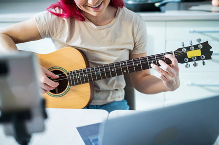 A woman sits in the kitchen during a remote acoustic guitar lesson. A girl learns to play the guitar and watches educational videos on a laptopの写真素材