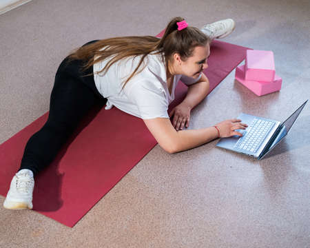 Young caucasian fat woman doing bends on a sports mat and watching a training video on a laptop. A...