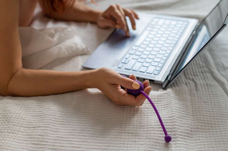 A woman lies in bed holding a vibrator and watching porn on a laptop