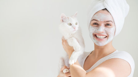 A woman with a towel on her hair and a clay mask on her face is holding a white fluffy cat on a...