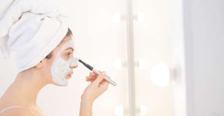 Beautiful caucasian woman with a towel on her hair applies a clay face mask. Taking care of beauty at homeの写真素材