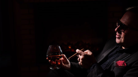 A mature man with glasses drinks brandy and smokes a cigar while sitting in an armchair by the fireplace in the dark. The concept of an elite gentlemens clubの写真素材