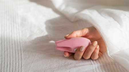 A faceless woman lies in bed under a blanket holding a clitoral vibrator. The girl masturbates