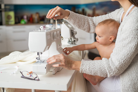 A young woman sews in the kitchen and holds a small child. Mom teaches her little son to sew on a...