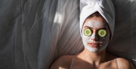 A woman with a towel on her hair and in a clay face mask and cucumbers in front of her eyes lies on the sheetの写真素材