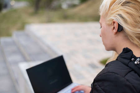 Blonde woman with hearing aid sits at laptop outdoors.の写真素材