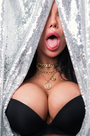 Portrait of a sexy brunette woman with big breasts dressed in a black bra peeking out from behind curtains of silver sequins.の写真素材