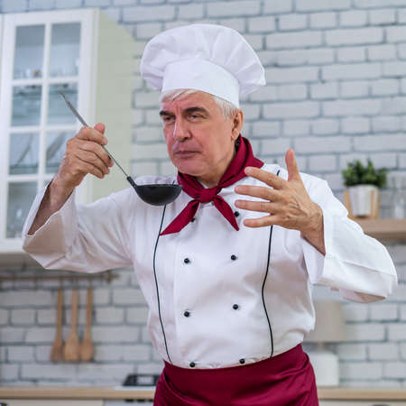 Portrait of smiling elderly chef tasting a scoop dish in the kitchen.の写真素材