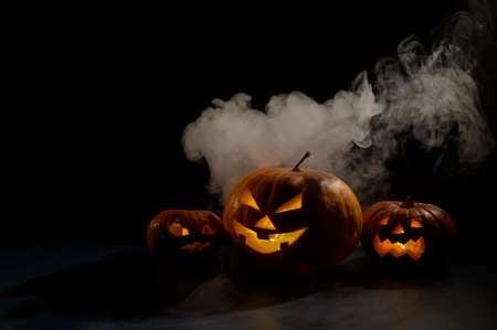 Three creepy halloween steaming pumpkins with a carved luminous smirk on a black background. A handmade jack-o-lantern head with a candle inside in the dark among the fog. Trick or treat. Postcard.の写真素材