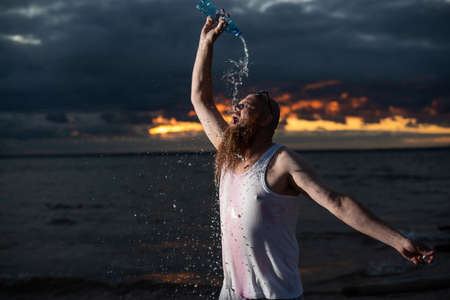 A humorous portrait of a brutal man pouring soda from a bottle on the beach at sunsetの写真素材