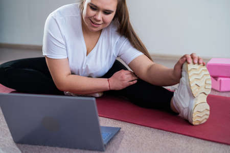 Young caucasian fat woman doing bends on a sports mat and watching a training video on a laptop. A...