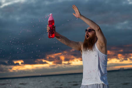 A humorous portrait of a brutal man pouring soda from a bottle on the beach at sunsetの写真素材