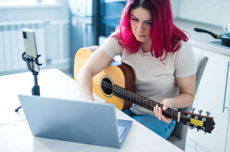 A woman sits in the kitchen during a remote acoustic guitar lesson. A girl learns to play the guitar and watches educational videos on a laptopの写真素材
