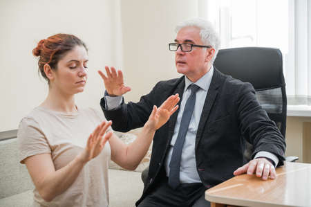 Mature gray-haired man hypnotizes Caucasian woman during hypnotherapy session. The psychologist uses...