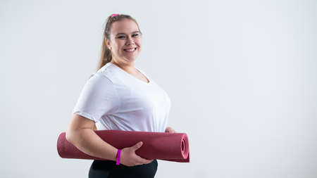 Young fat caucasian woman holding a sport mat. Charming plus size model in sportswear stands on a...