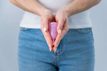 A faceless woman wearing jeans and a white T-shirt holds a pink menstrual cup on a white background. Great alternative to tompons and padsの写真素材