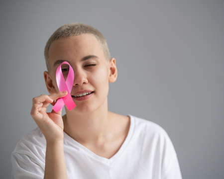 Young woman with short hair wearing a white t-shirt holding a pink ribbon as a symbol of breast cancer on a white background.の写真素材