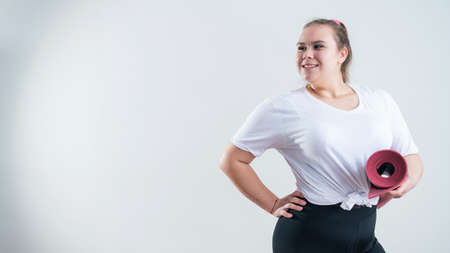Young fat caucasian woman holding a sport mat. Charming plus size model in sportswear stands on a white backgroundの写真素材