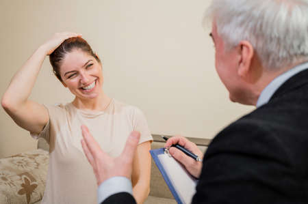 Mature gray-haired male psychotherapist with clipboard in session with female patient. Smiling caucasian woman at a psychologist appointment.の写真素材
