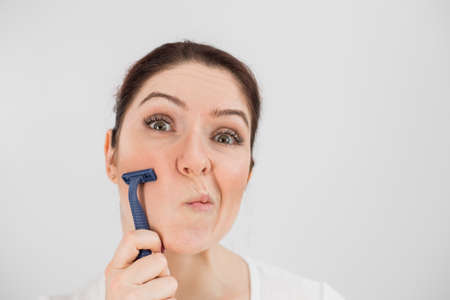 Caucasian funny woman shaves her face with a straight razor on a white background. Copy space.の写真素材