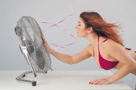 Red-haired Caucasian woman in bikini enjoys cold airflow from electric fan against white background. Climate control on a hot summer dayの写真素材