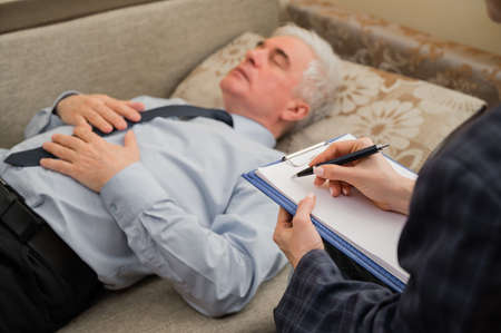 An elderly man lies on a couch during an appointment with a psychotherapist. An elderly male patient in an individual consultation with a female psychologistの写真素材