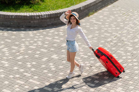 Happy caucasian young woman in hat and shorts holding a large red suitcase at an open ageの写真素材
