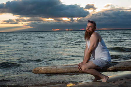 Funny bald man with red beard posing on the beach at sunset. A humorous male parody of a glamorous girl.の写真素材