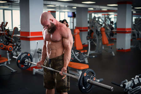 Bald man with a beard in the gym. Muscular bodybuilder guy doing barbell exercises. Strong man with a naked torso. The young athlete is preparing for weightlifting competitions.の写真素材
