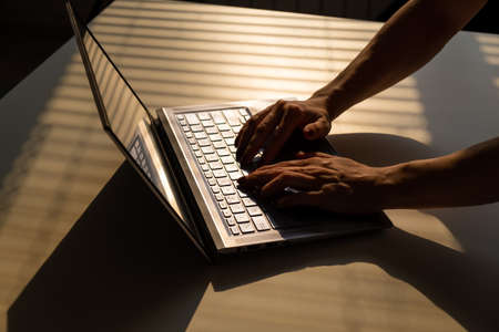 A woman is typing on a laptop keyboard on a white table. The shadow from the blinds falls on the...